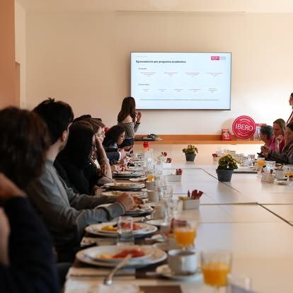 Asistentes sentados a lo largo de una mesa durante una reunión con presentación en pantalla