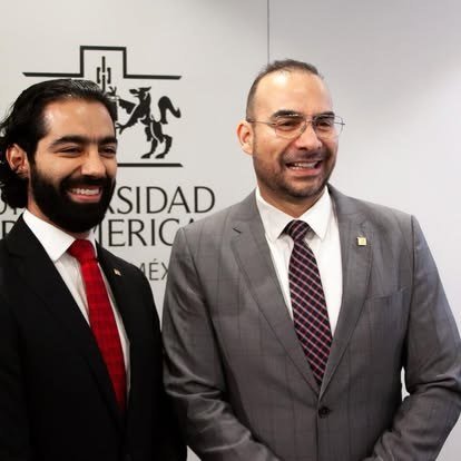 Dos egresados posan frente al escudo de la Universidad Iberoamericana